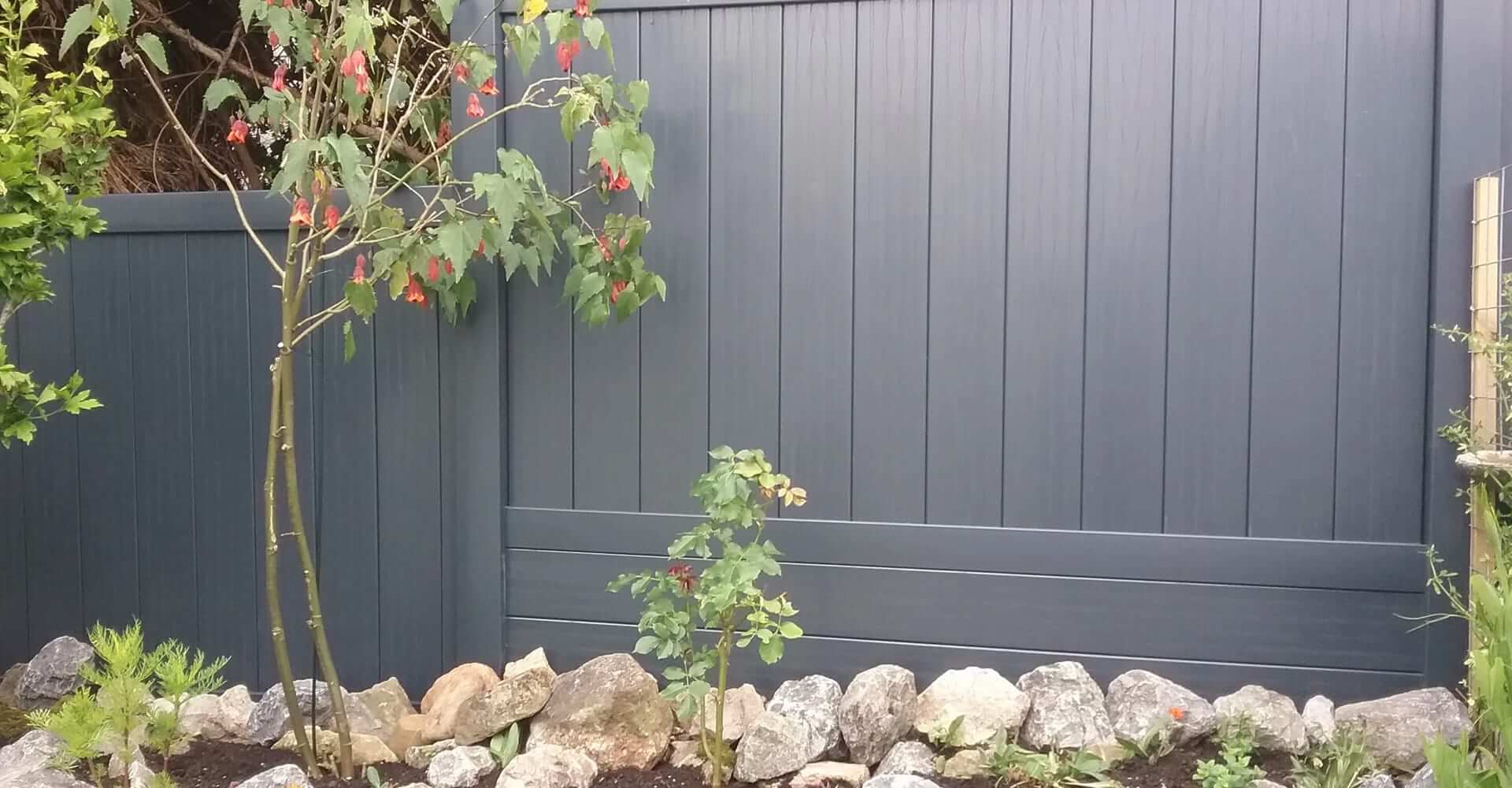 Dark gray uPVC fencing with plants growing against it and garden rocks below